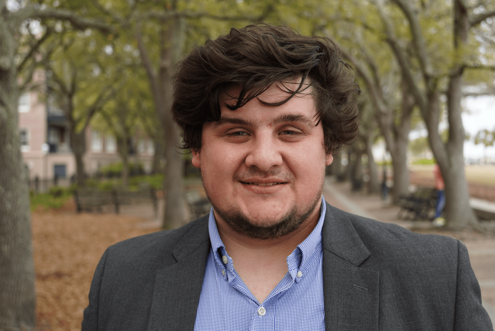 A man with wavy dark hair and a short beard, wearing a gray blazer and blue checkered shirt, smiles outdoors on a tree-lined path with benches and leaves on the ground.