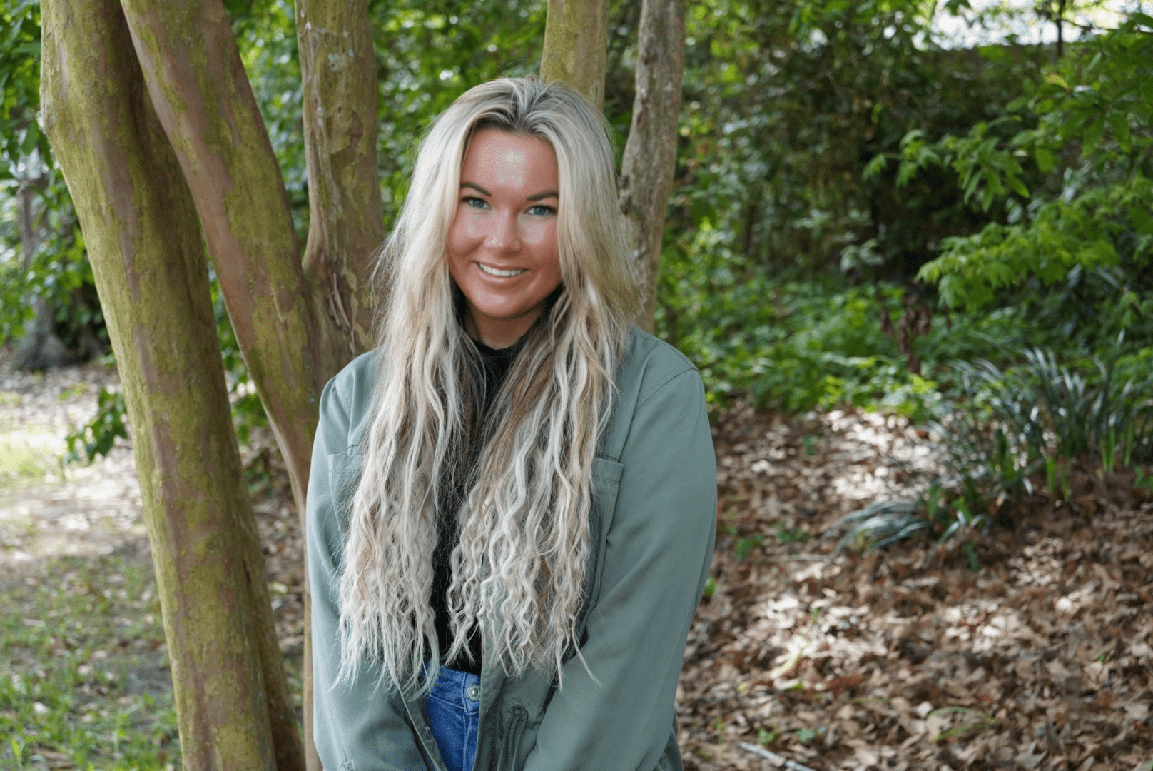 A woman with long, wavy blonde hair smiles while standing outdoors in front of trees and greenery. She is wearing a green jacket over a black top and blue jeans.