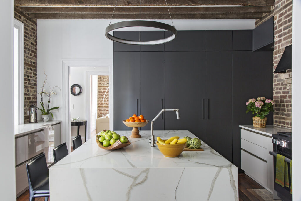 A modern kitchen with a marble island, black cabinets, exposed brick walls, fruit bowls with bananas, apples, and oranges, and a round pendant light hanging above. Orchids and a potted plant add greenery to the space.