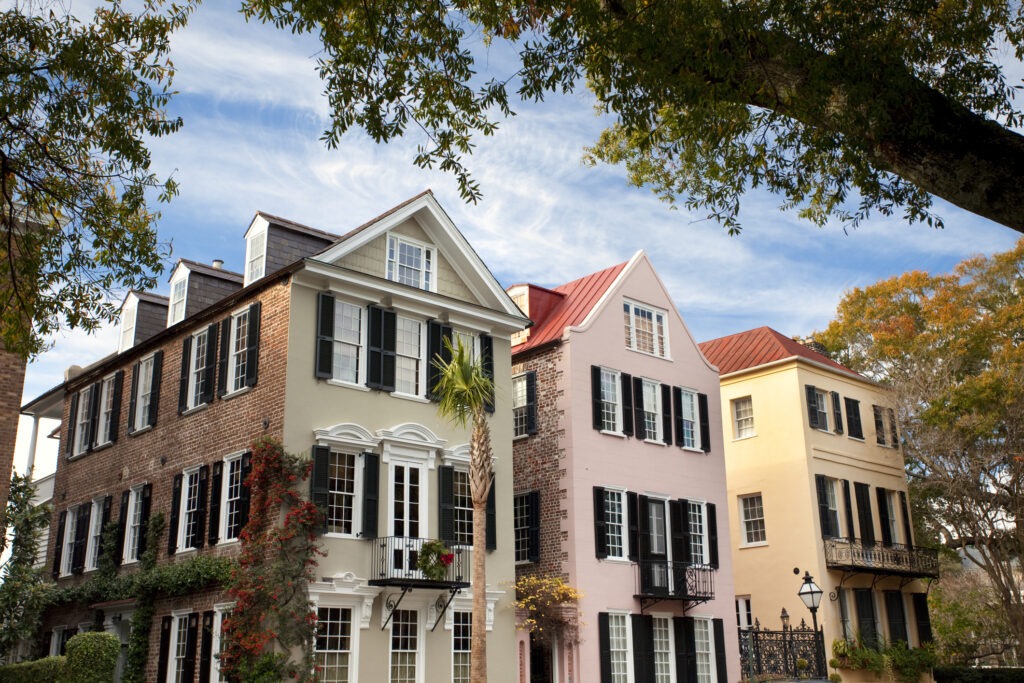 Three colorful historic row houses with black shutters and balconies stand side by side under a partly cloudy sky, framed by leafy tree branches in the foreground.