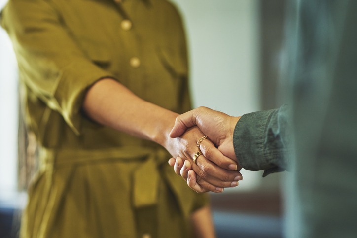 Two people shake hands, one wearing a green dress and the other a dark green shirt. The focus is on their handshake, symbolizing agreement or partnership, with a blurred background.