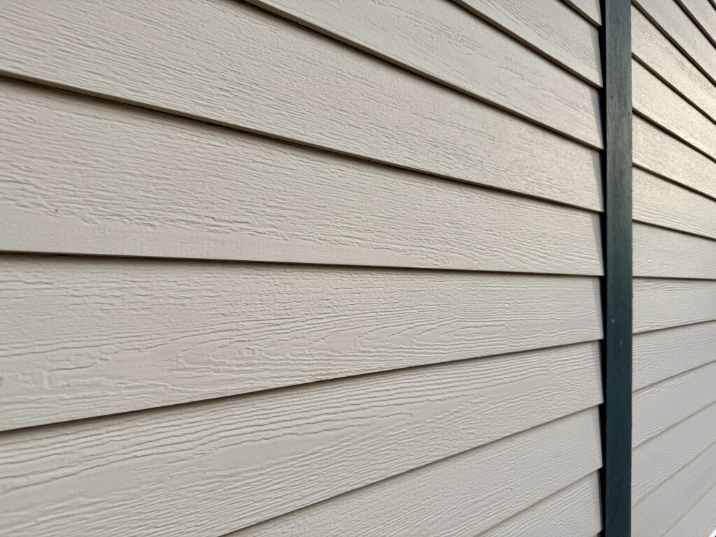 Close-up view of beige horizontal vinyl siding on the exterior of a building, with a vertical dark trim piece on the right side. The siding features a wood grain texture.