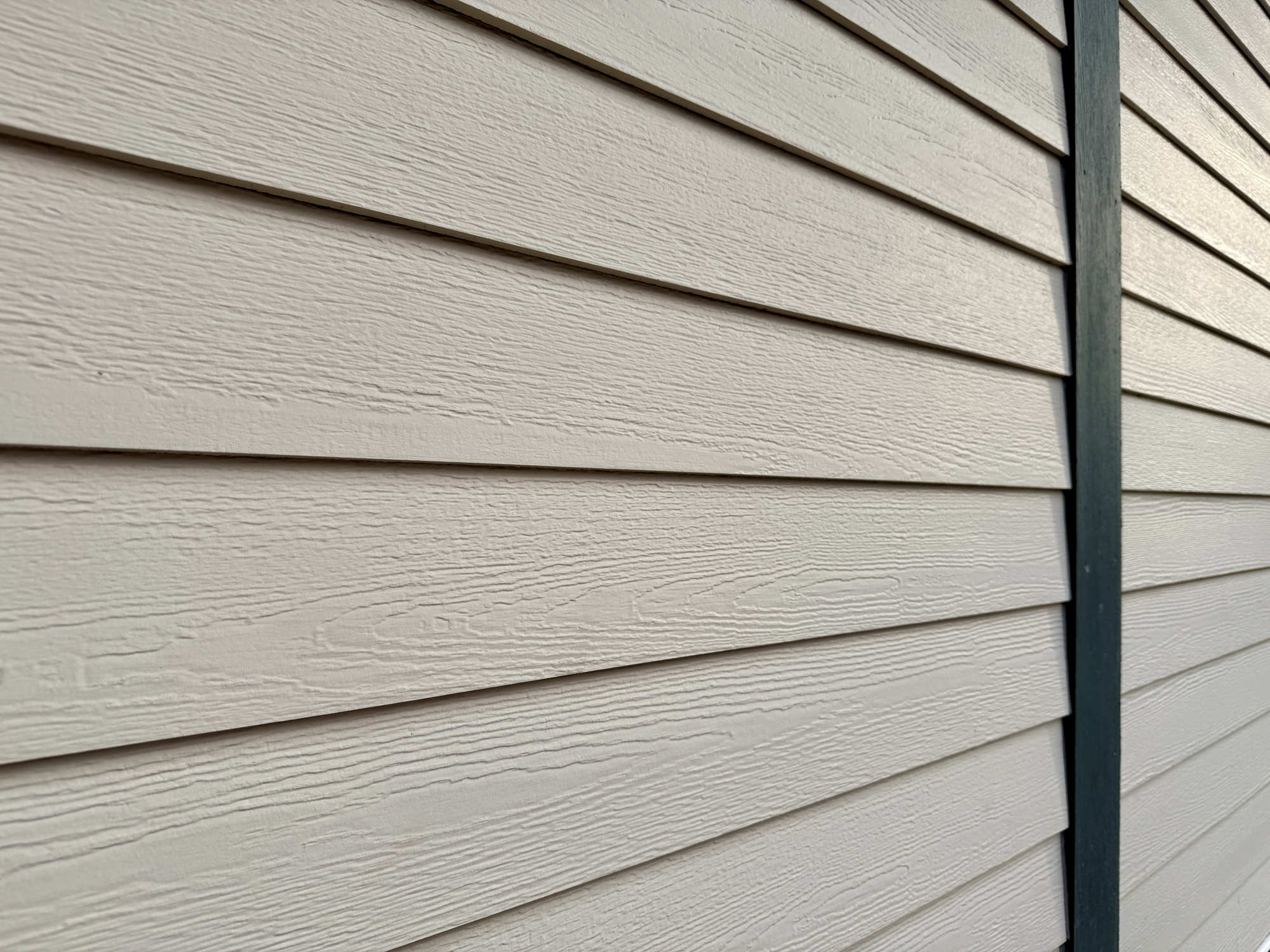 Close-up view of beige horizontal vinyl siding on the exterior of a building, with a vertical dark trim piece on the right side. The siding features a wood grain texture.