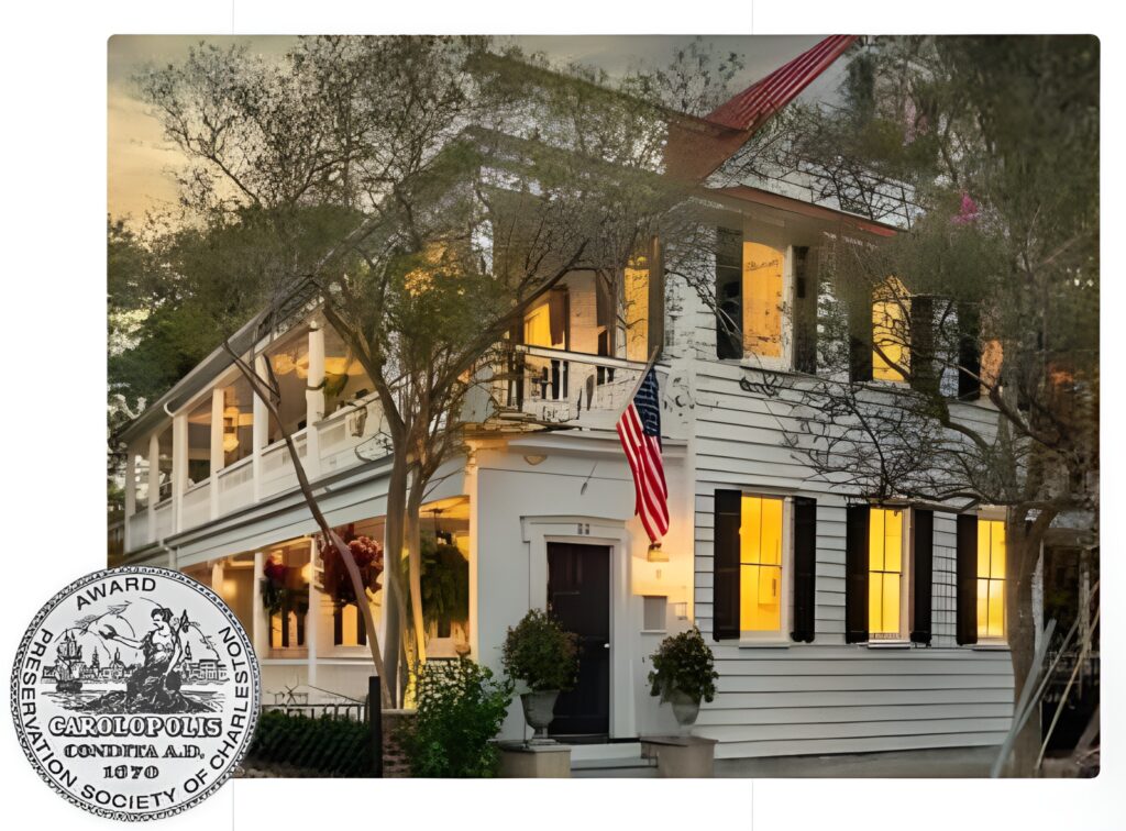 A two-story white house with black shutters is lit warmly from within. An American flag hangs by the front door, and trees partly frame the scene. A round award seal appears in the bottom left corner.
